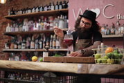 Een barman met hoed en baard maakt een drankje klaar in een groot wijnglas. Op de achtergrond zijn planken met verschillende flessen sterke drank te zien. Het perspectief is recht op de barman gericht.