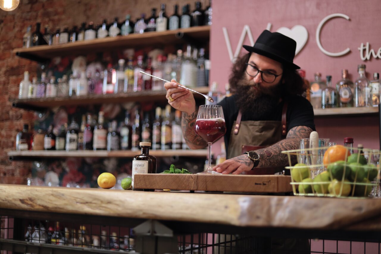 Een barman met hoed en baard maakt een drankje klaar in een groot wijnglas. Op de achtergrond zijn planken met verschillende flessen sterke drank te zien. Het perspectief is recht op de barman gericht.