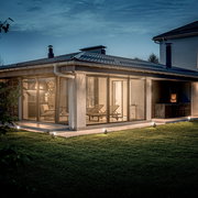 Moderne Terrasse mit Glaswänden und gemütlichen Liegestühlen, beleuchtet von Bodenspikes in der Abenddämmerung, seitliche Perspektive.