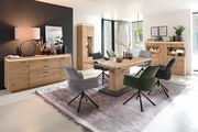 Wohnzimmer mit modernem Esstisch-Set aus Holz, bestehend aus einem Esstisch, sechs Stühlen in Grau und Grün, einem Sideboard, einer Vitrine und einem Wandboard. Perspektive: Frontalansicht des gesamten Sets in einem hellen Raum mit großen Fenstern.