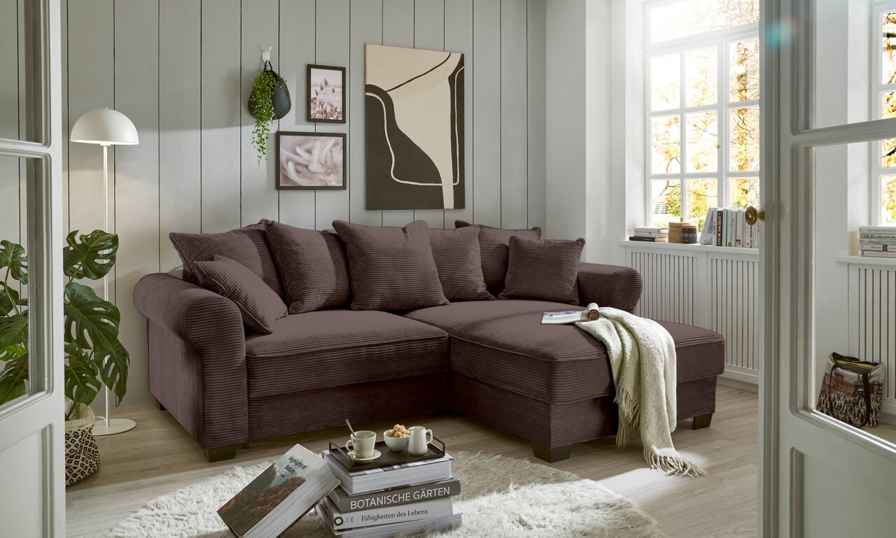 Hom´in Ecksofa MAUREEN Gemütliche braune Rundecke in einem hellen Wohnzimmer mit großen Fenstern, dekoriert mit Pflanzen und Kunstwerken, aus der Perspektive von vorne rechts.