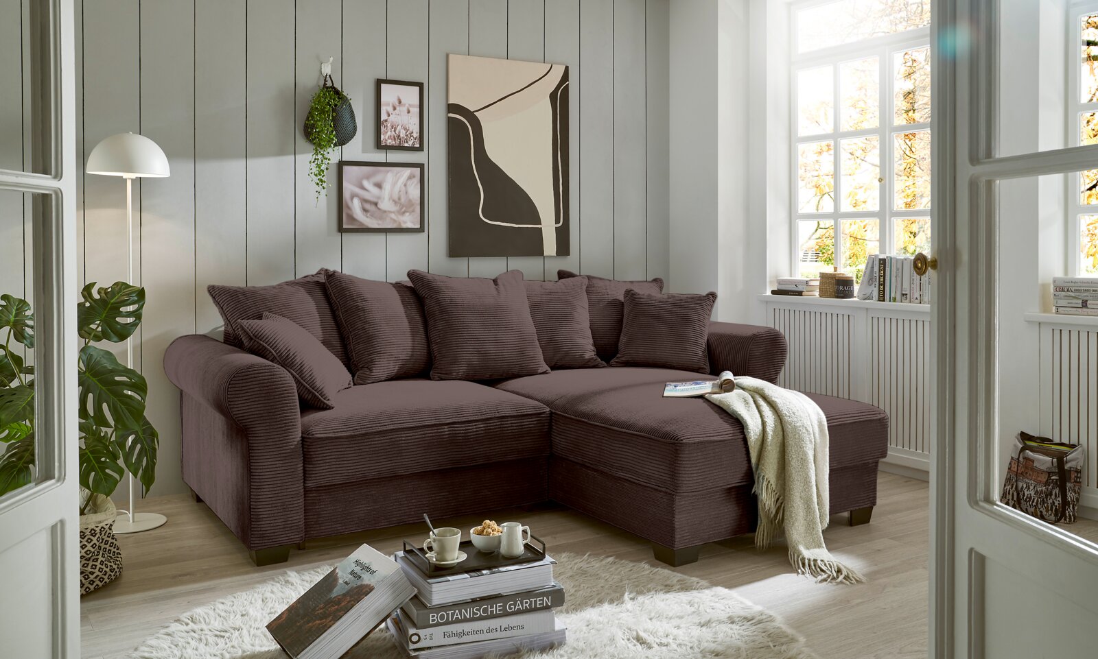 Gemütliche braune Rundecke in einem hellen Wohnzimmer mit großen Fenstern, dekoriert mit Pflanzen und Kunstwerken, aus der Perspektive von vorne rechts.