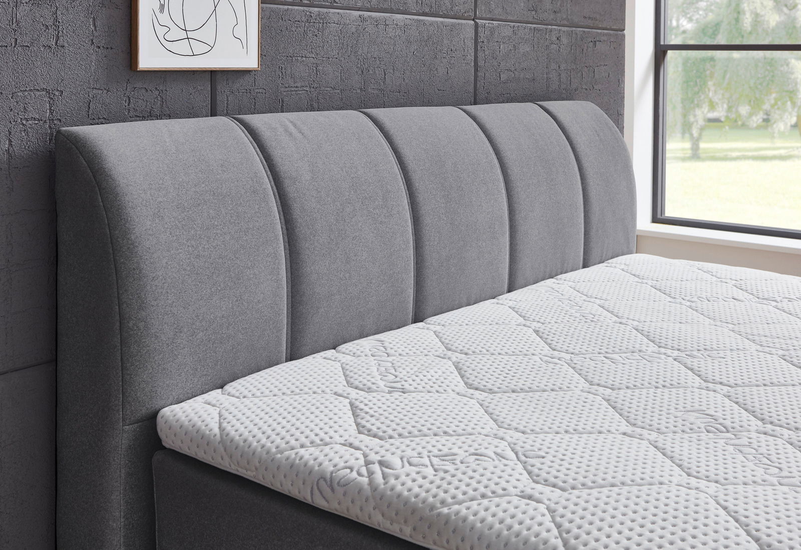 Seitliche Ansicht eines grauen Boxspringbetts mit gepolstertem Kopfteil und weißer Matratze in einem modernen Schlafzimmer.