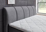 Seitliche Ansicht eines grauen Boxspringbetts mit gepolstertem Kopfteil und weißer Matratze in einem modernen Schlafzimmer.