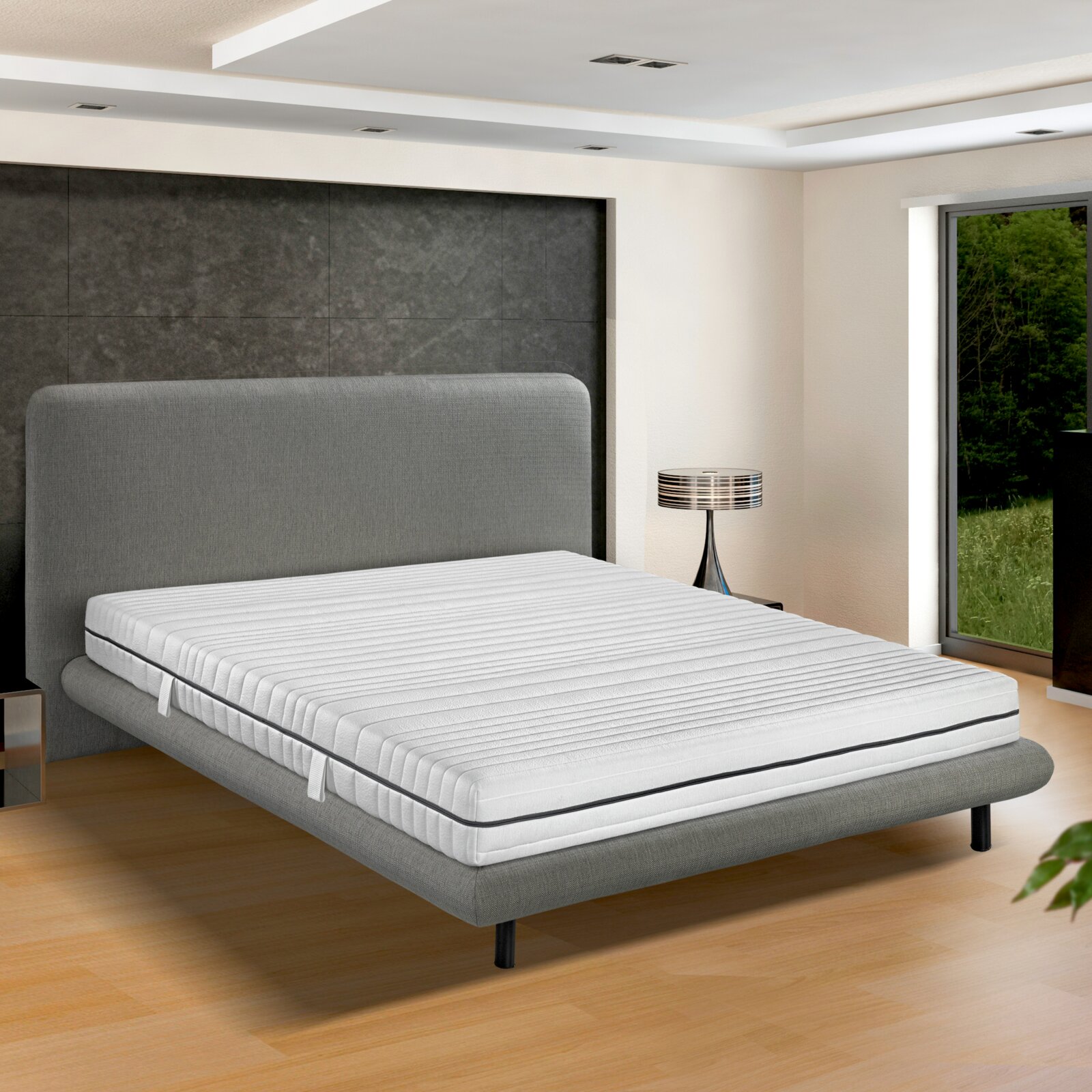 Seitliche Perspektive auf eine moderne TTFK-Matratze H3 auf einem grauen Bettgestell in einem stilvollen Schlafzimmer mit großen Fenstern.