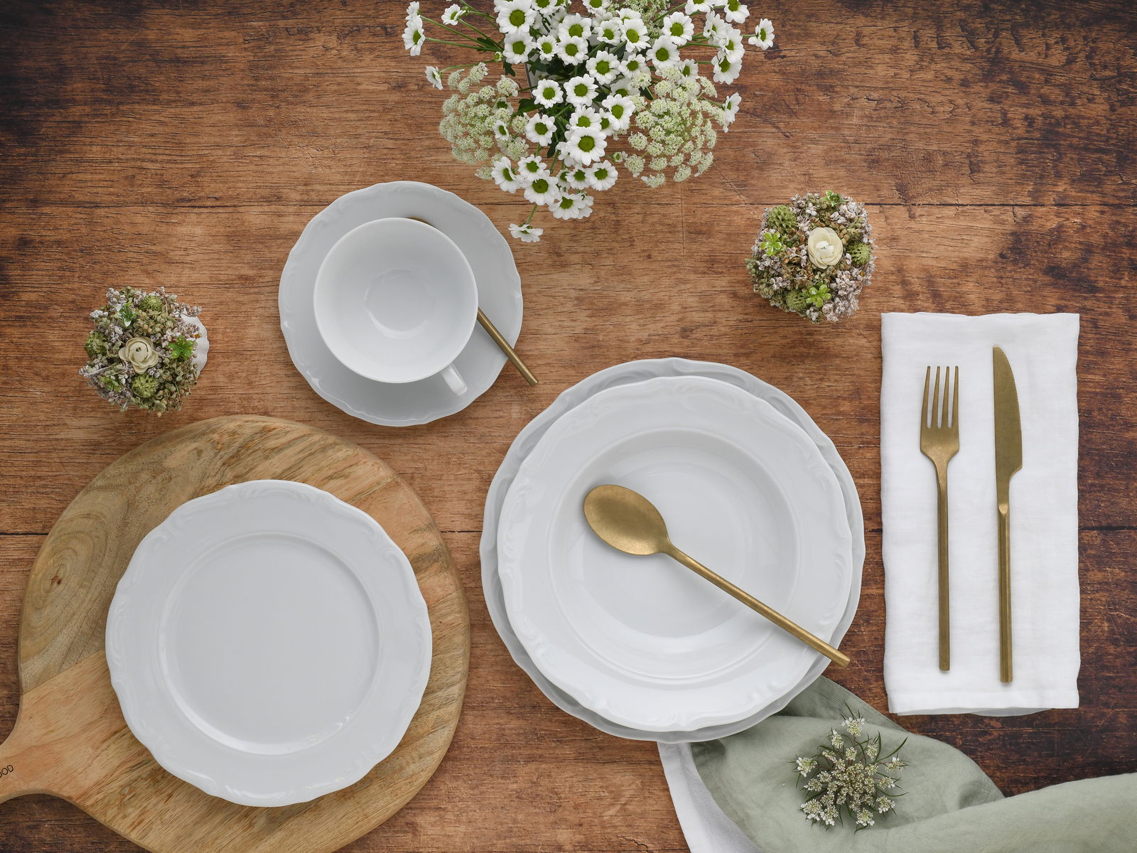 Vogelperspektive auf ein elegantes Kombiservice-Set Maria Theresia mit weißen Tellern, Tasse und goldfarbenem Besteck, dekoriert mit Blumen auf einem Holztisch.