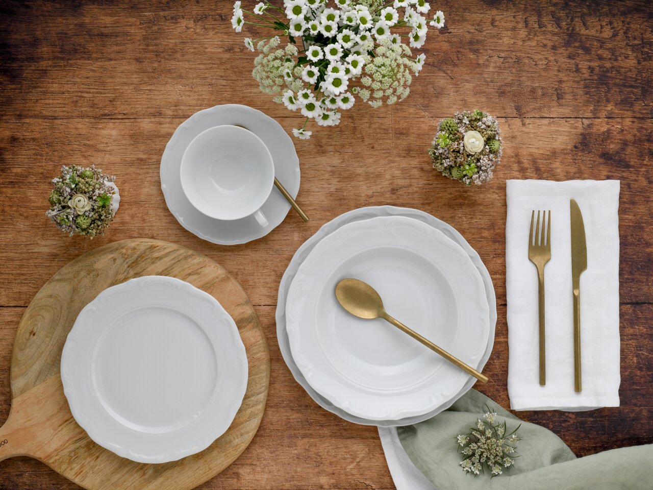 Vogelvluchtperspectief op een elegante servieset Maria Theresia met witte borden, kopjes en goudkleurig bestek, versierd met bloemen op een houten tafel.