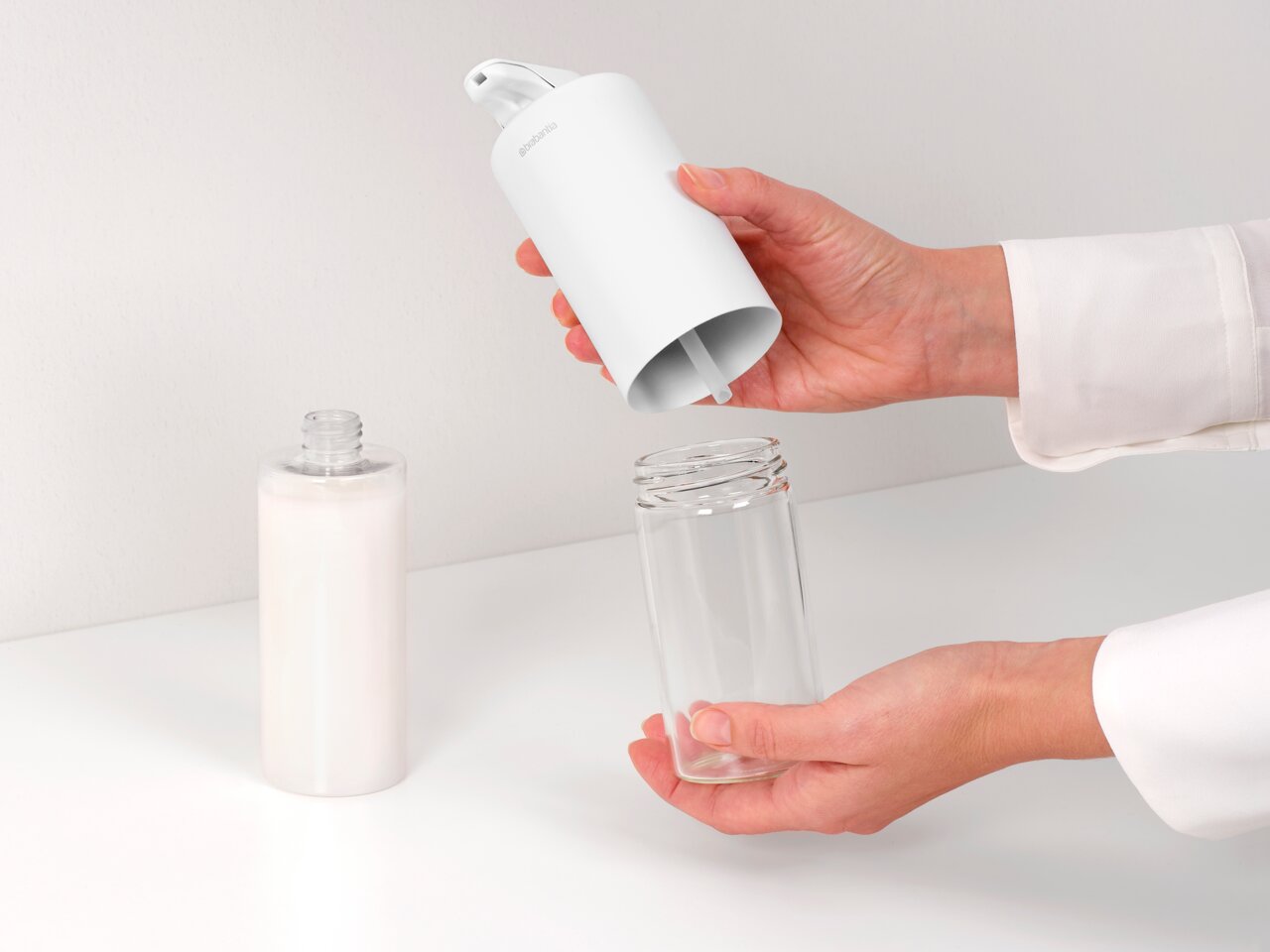Close-up van een witte Brabantia SINKSTYLE-zeepdispenser die door iemand wordt vastgehouden. De dispenser wordt boven een doorzichtige fles gehouden om deze bij te vullen met zeep.