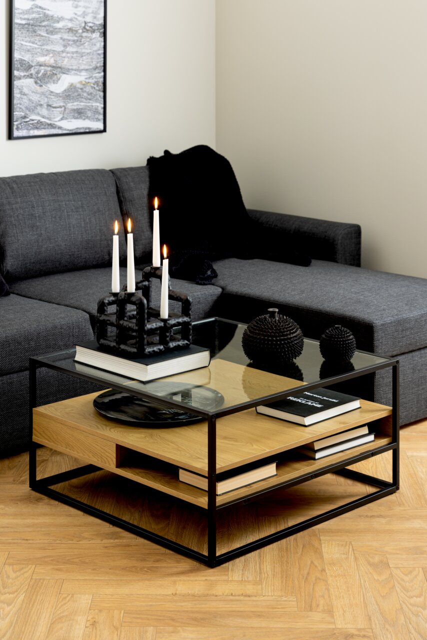 Moderner Couchtisch mit Glasplatte und Holzablage, dekoriert mit Kerzen und Büchern, aus seitlicher Perspektive fotografiert.