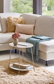 Helles, strukturiertes Ecksofa mit Kissen, Decke und rundem Beistelltisch im modernen Wohnzimmer vor großen Fenstern, Perspektive: leicht seitliche Vorderansicht