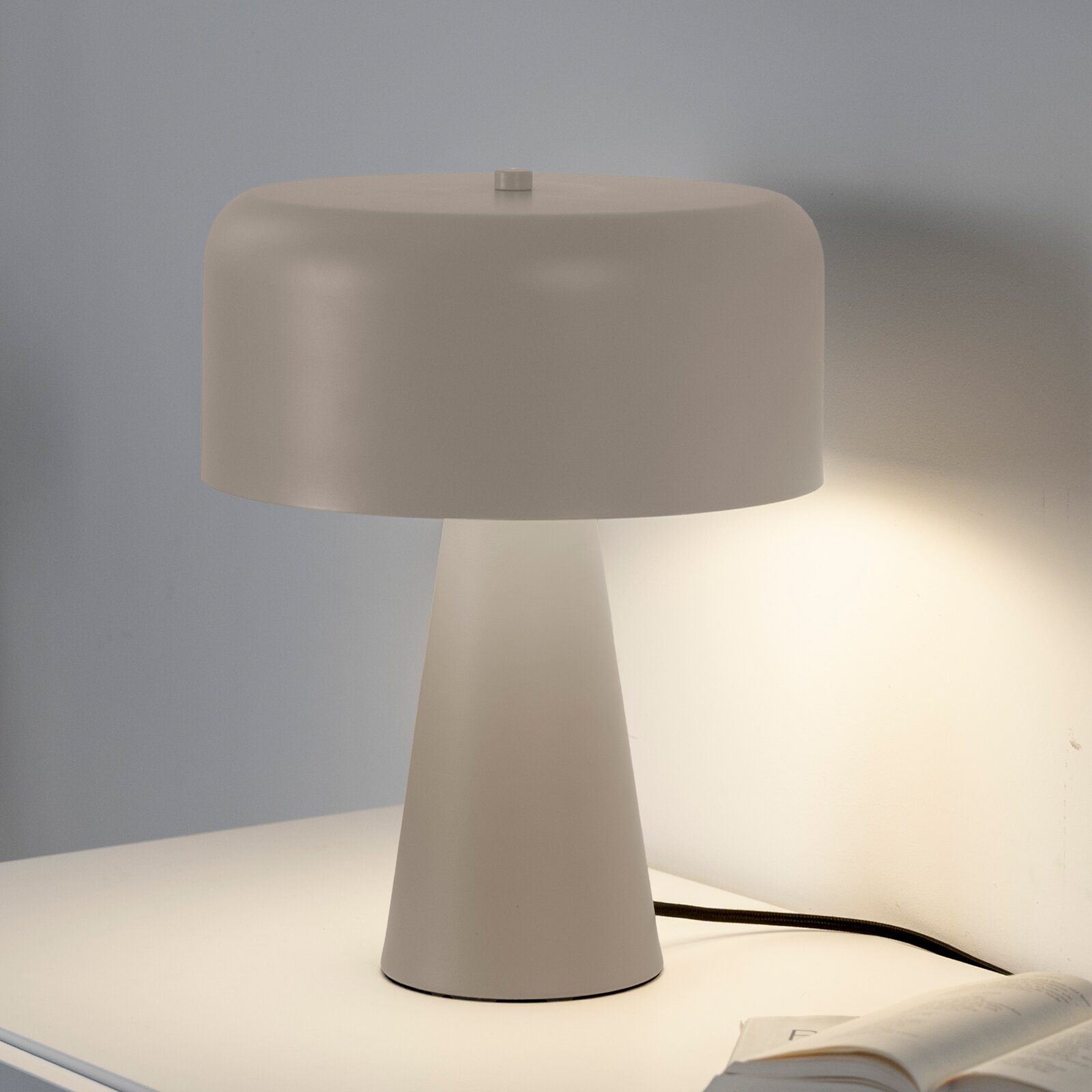 Weiße Tischlampe mit breitem, abgerundetem Lampenschirm und konischem Fuß auf einem Tisch, eingeschaltet; Frontansicht leicht von links.