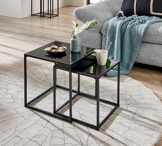 Zwei ineinander schiebbare Beistelltische im Set mit schwarzer Glasplatte und schwarzem Metallgestell auf hellem Teppich vor grauem Sofa, perspektivisch in schräger Seitenansicht von oben fotografiert.