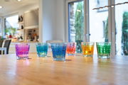 Sechs bunte Trinkgläser, darunter ein violettes Glas, auf einem Holztisch in einer modernen Küche, aus seitlicher Perspektive fotografiert.