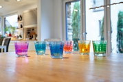 Sechs bunte Trinkgläser, darunter ein violettes Glas, auf einem Holztisch in einer modernen Küche, aus seitlicher Perspektive fotografiert.