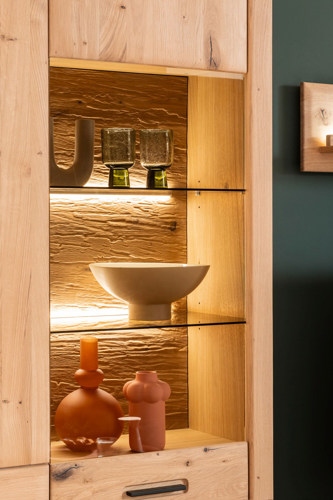 Detailansicht einer beleuchteten Holzwandkombination mit Glasregalen, dekoriert mit modernen Vasen und Schalen in warmen Erdtönen.