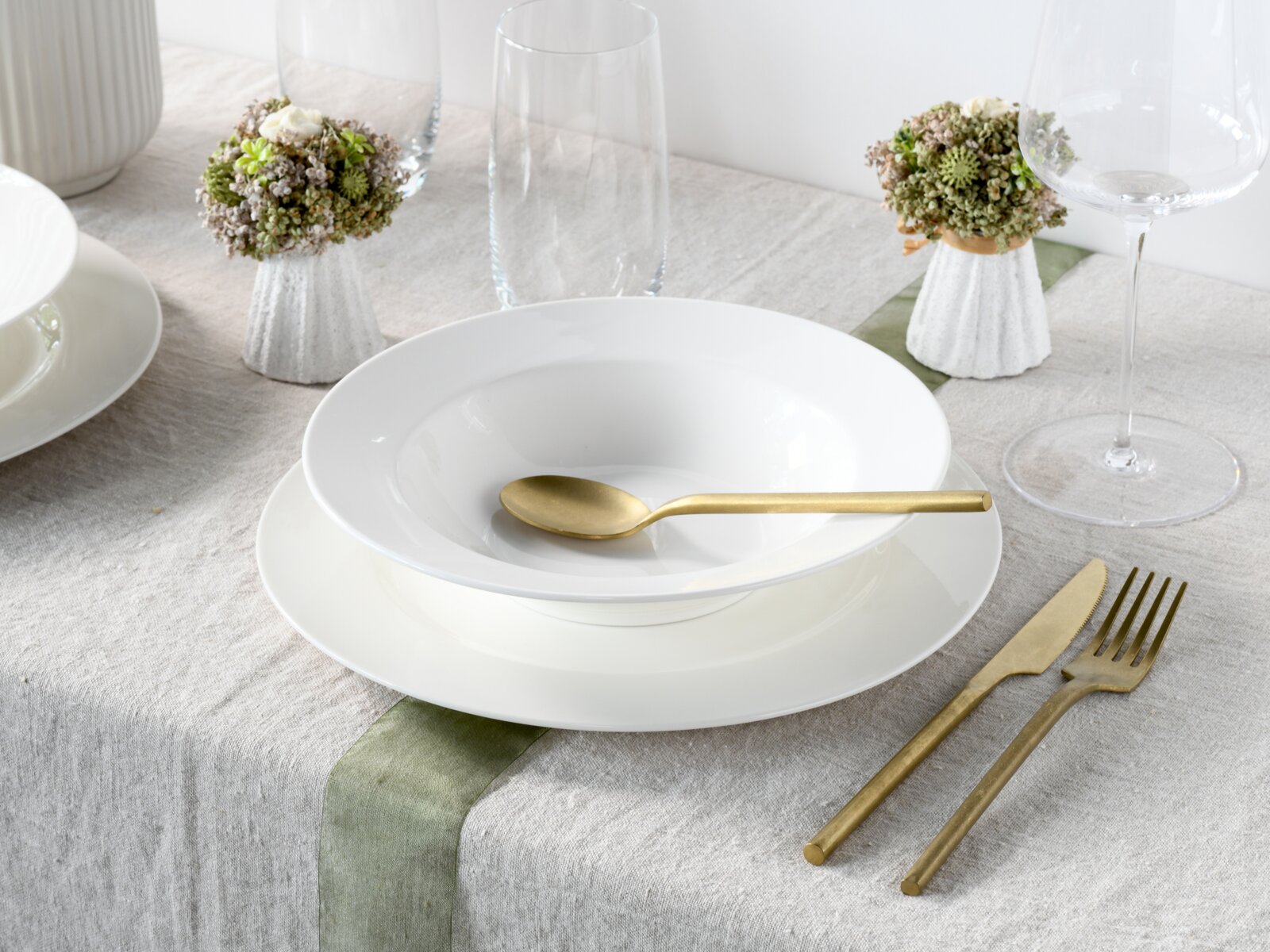 Modern gedeckter Tisch mit weißem Porzellanteller und tiefer Schale, goldfarbenem Besteck und Gläsern auf Leinentischdecke; Perspektive: leicht schräg von oben, frontal auf das Gedeck