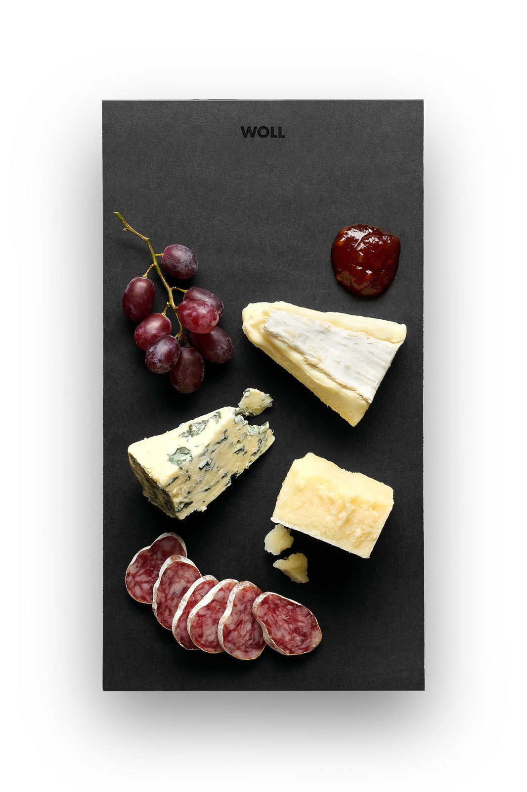Vogelperspektive auf ein schwarzes Servierbrett mit verschiedenen Käsesorten, Trauben, Salami und einem Klecks Marmelade.