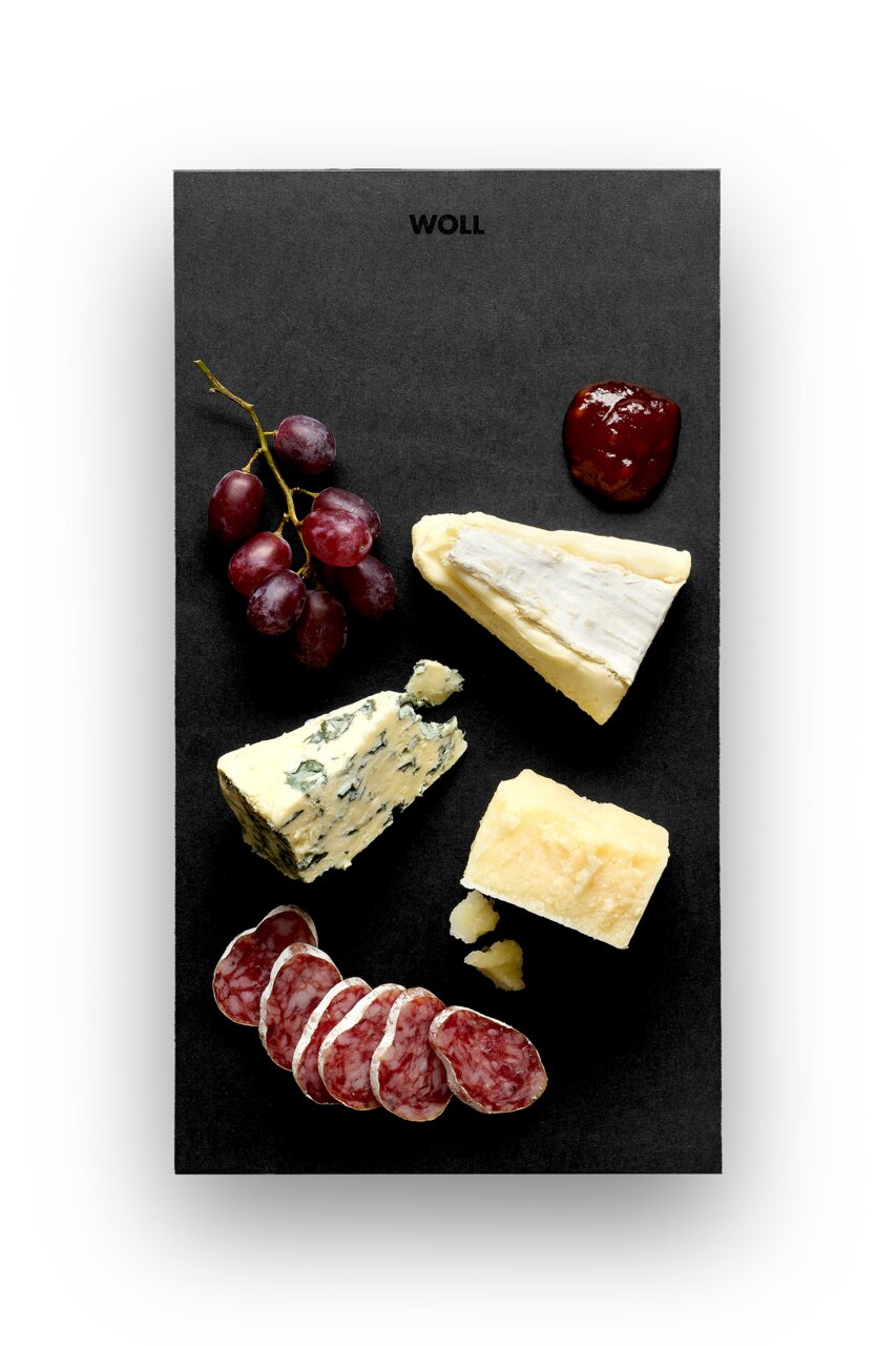 Vogelperspectief op een zwarte serveerschaal met verschillende kaassoorten, druiven, salami en een klodder jam.