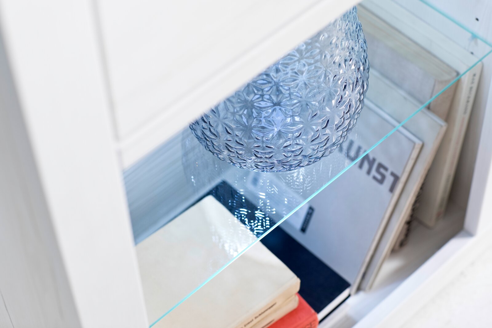 Detailfoto schuin van bovenaf: blauwe, gestructureerde glazen pot op een glazen plank in een witte boekenkast, met daarnaast liggende boeken