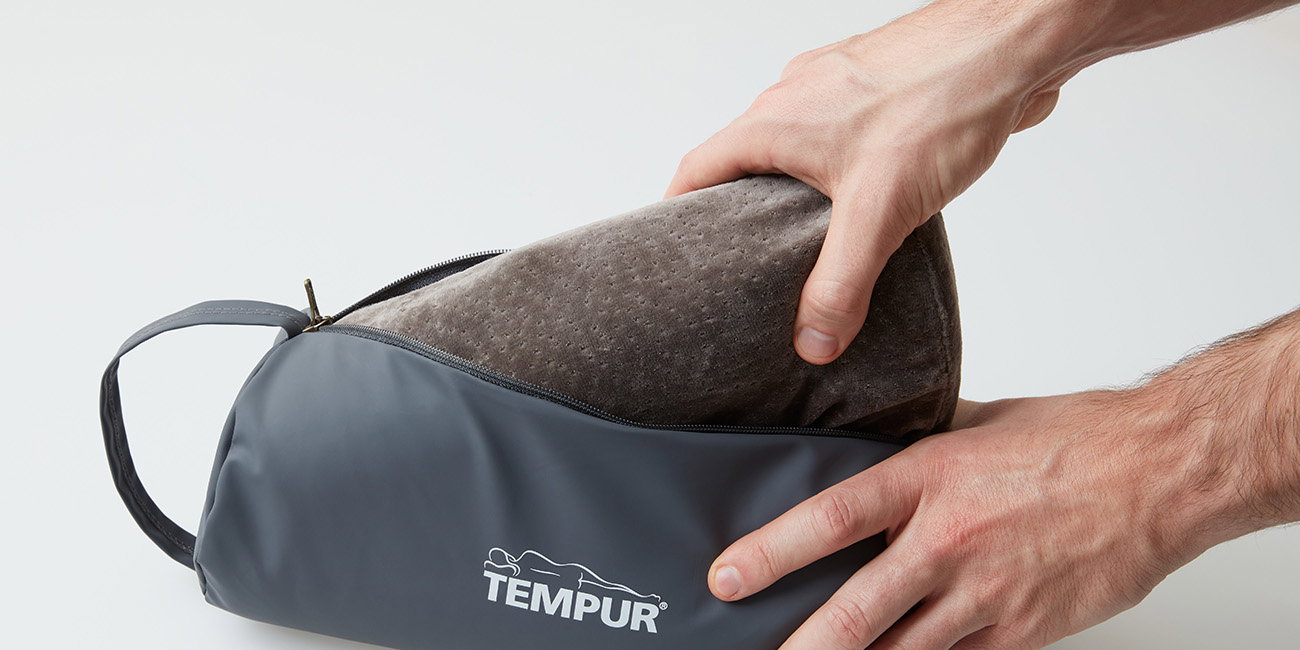 Hände verstauen ein Reisekissen in einer grauen Tasche mit Tempur-Logo.