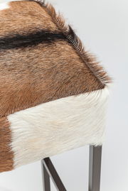 Nahaufnahme eines Hockers mit Fellbezug in Braun- und Weißtönen, seitliche Perspektive