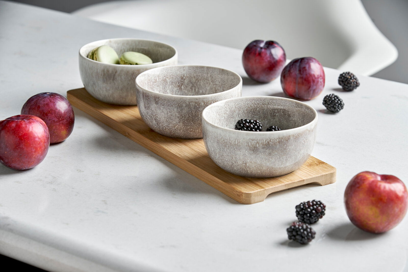 Dreiteiliges Schalen-Set auf einem Holztablett, gefüllt mit Macarons und Brombeeren, umgeben von Pflaumen, seitliche Perspektive.