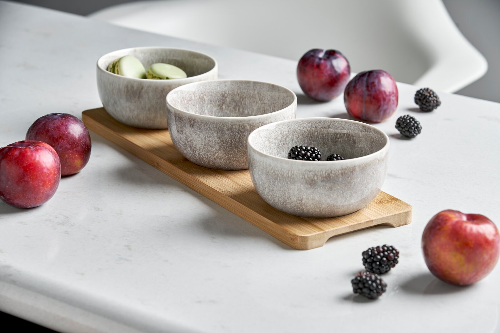 Dreiteiliges Schalen-Set auf einem Holztablett, gefüllt mit Macarons und Brombeeren, umgeben von Pflaumen, seitliche Perspektive.