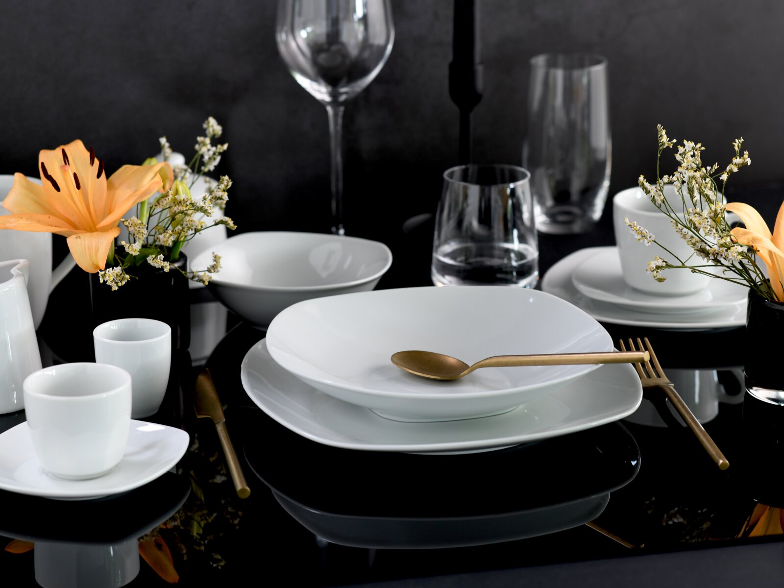 Modern gedeckter Tisch mit weißem Porzellan-Geschirr (Teller und Schalen), weißen Tassen, Glaswaren und goldfarbenem Besteck auf schwarzem Tisch, dekoriert mit orangefarbenen Lilien und kleinen weißen Blüten; Perspektive: leicht von vorne/seitlich in Auge