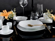 Modern gedeckter Tisch mit weißem Porzellan-Geschirr (Teller und Schalen), weißen Tassen, Glaswaren und goldfarbenem Besteck auf schwarzem Tisch, dekoriert mit orangefarbenen Lilien und kleinen weißen Blüten; Perspektive: leicht von vorne/seitlich in Auge
