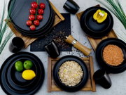 Vogelperspektive auf ein stilvolles, schwarzes Kombiservice-Set mit Tellern, Schalen und Tassen, dekoriert mit frischen Zutaten wie Tomaten, Paprika, Linsen, Haferflocken, Limetten und Zitrone auf Holztabletts.