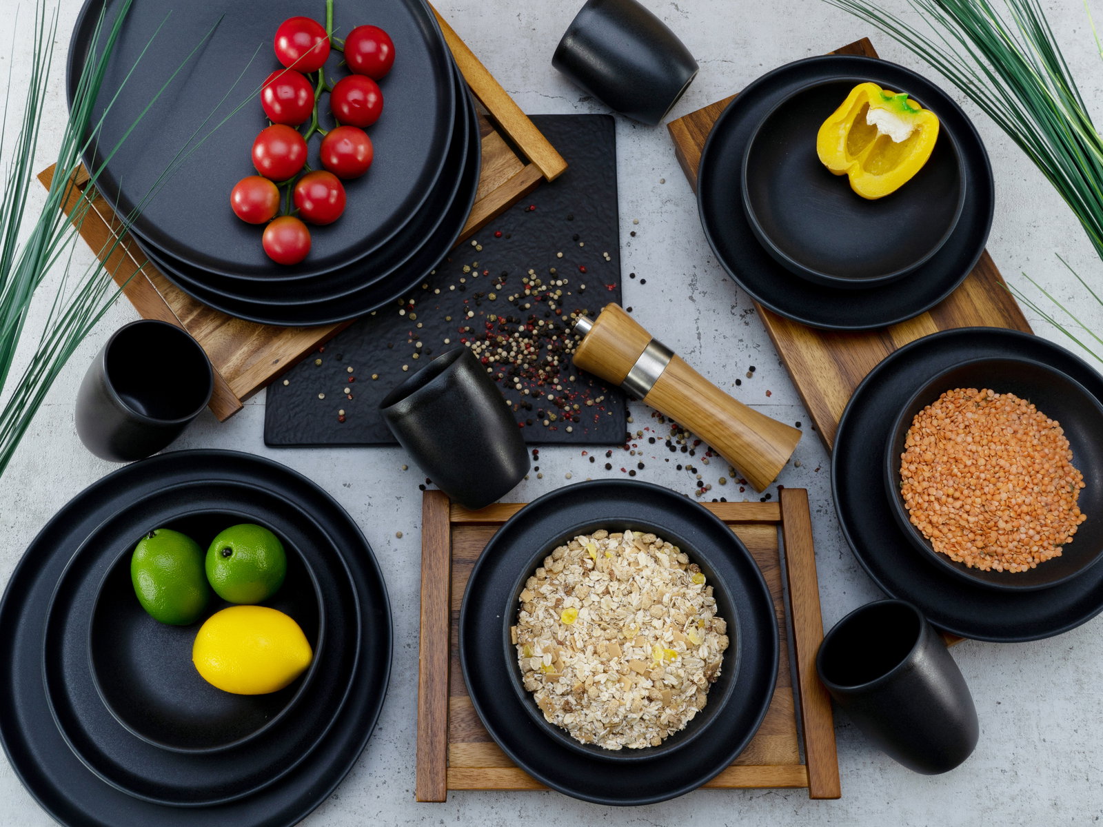 Vogelperspektive auf ein stilvolles, schwarzes Kombiservice-Set mit Tellern, Schalen und Tassen, dekoriert mit frischen Zutaten wie Tomaten, Paprika, Linsen, Haferflocken, Limetten und Zitrone auf Holztabletts.