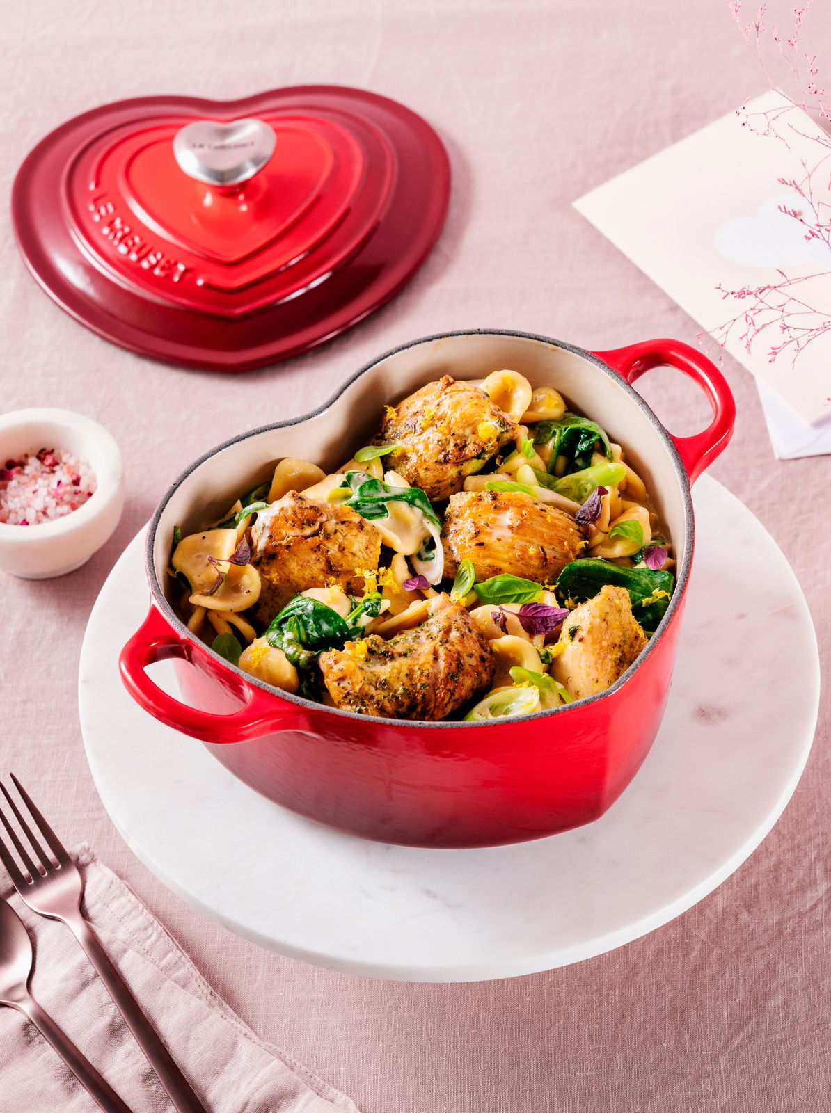 Herzförmiger roter Bräter mit Henkeln, gefüllt mit einem leckeren Gericht, auf einem Tisch von oben fotografiert.