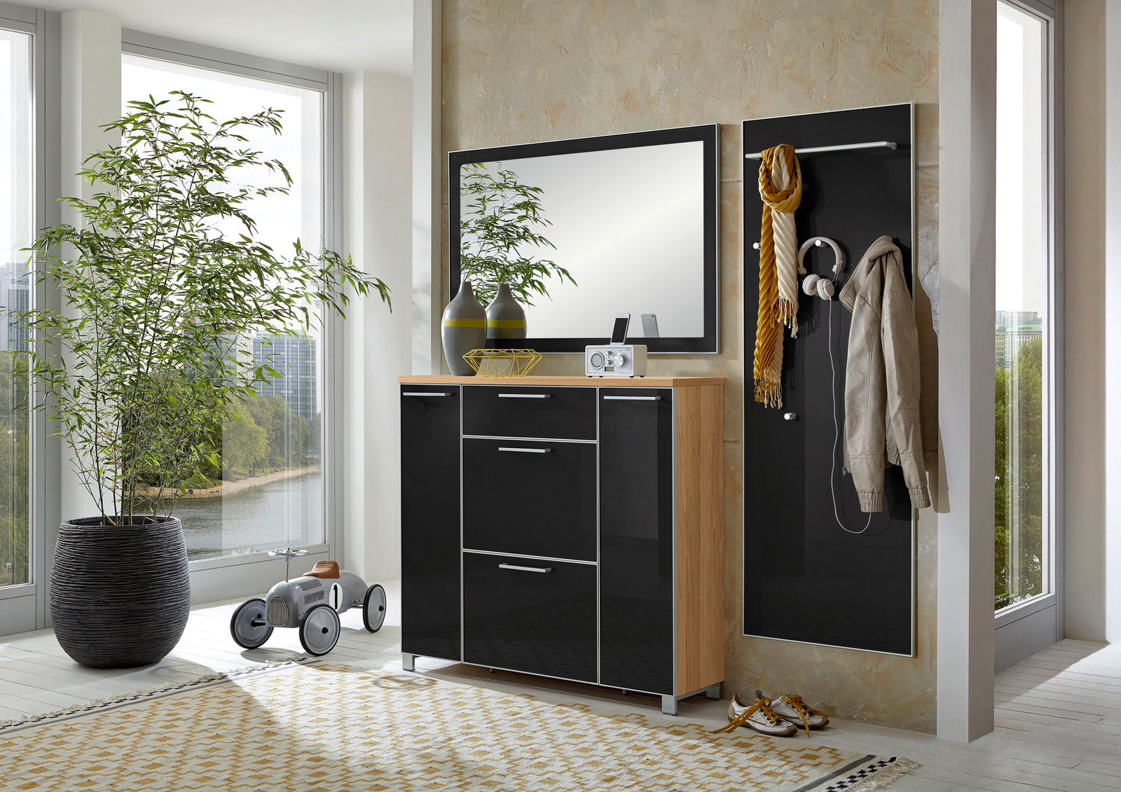 Modernes Garderoben-Set in einem hellen Raum mit großen Fenstern, bestehend aus einem schwarzen Schrank mit Holzdetails, einem Spiegel und einer Wandpaneele mit Haken. Perspektive von vorne.