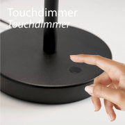 Nahaufnahme aus leicht schräger Vorderansicht: schwarzer Lampenfuß mit Touchdimmer-Taste, ein Finger berührt den Dimmer.