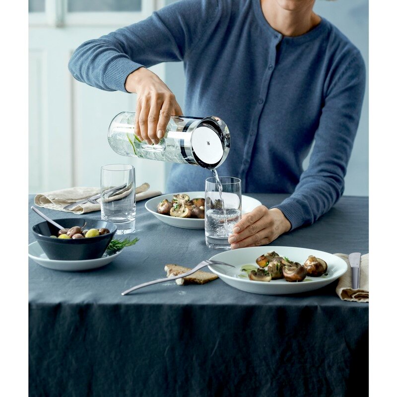 Person gießt Wasser aus einer eleganten Wasserkaraffe in ein Glas auf einem gedeckten Tisch, seitliche Perspektive.