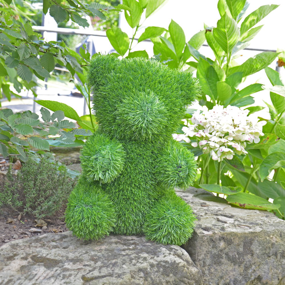 Grüner Bär aus Gras, frontal im Garten auf einem Stein sitzend, umgeben von Pflanzen.