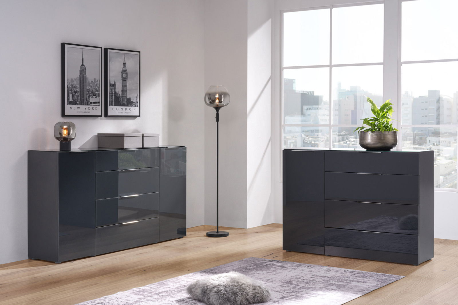 Moderne, dunkelgraue Kombikommode in einem hellen Raum mit großen Fenstern, dekoriert mit Pflanzen und Bildern, seitliche Perspektive.