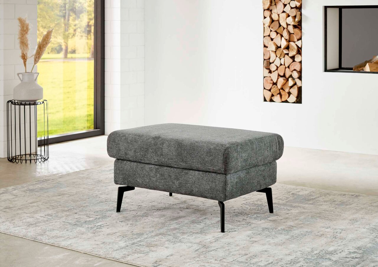 Grauer XL Hocker mit schwarzen Beinen in einem modernen Wohnzimmer, seitliche Perspektive.