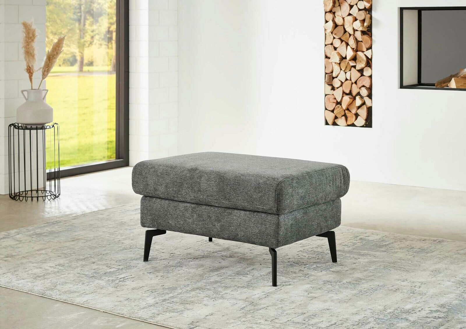 Grauer XL Hocker mit schwarzen Beinen in einem modernen Wohnzimmer, seitliche Perspektive.