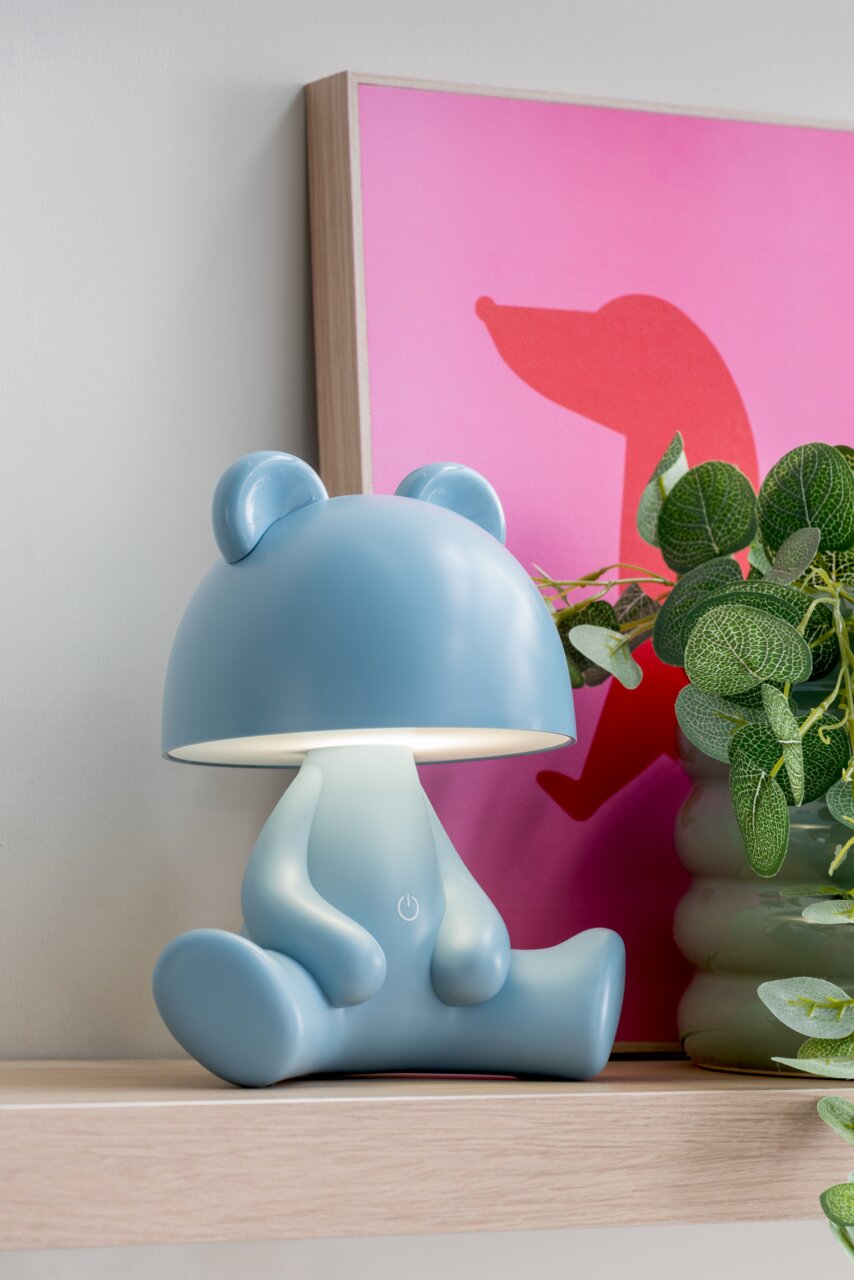 Blaue LED-Tischlampe in Bärenform, seitliche Perspektive, auf einem Regal neben einer Pflanze und einem pinken Bild.