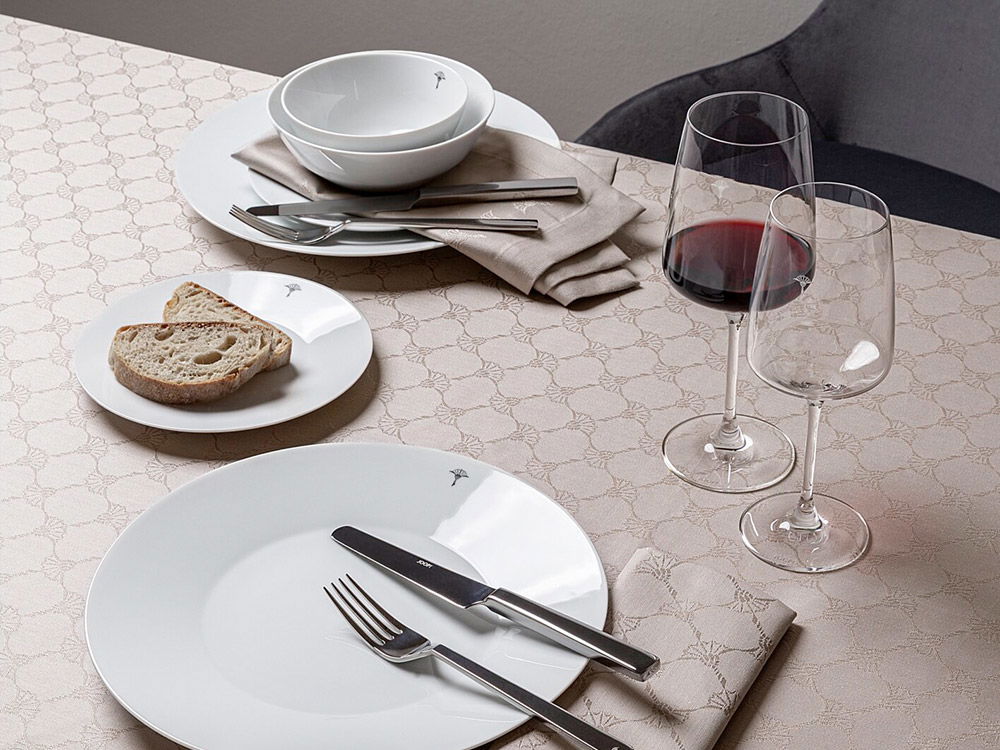 Essgeschirr mit Tellern, Besteck, Weingläsern, einer Serviette und einer Scheibe Brot auf einem strukturierten Tischtuch.