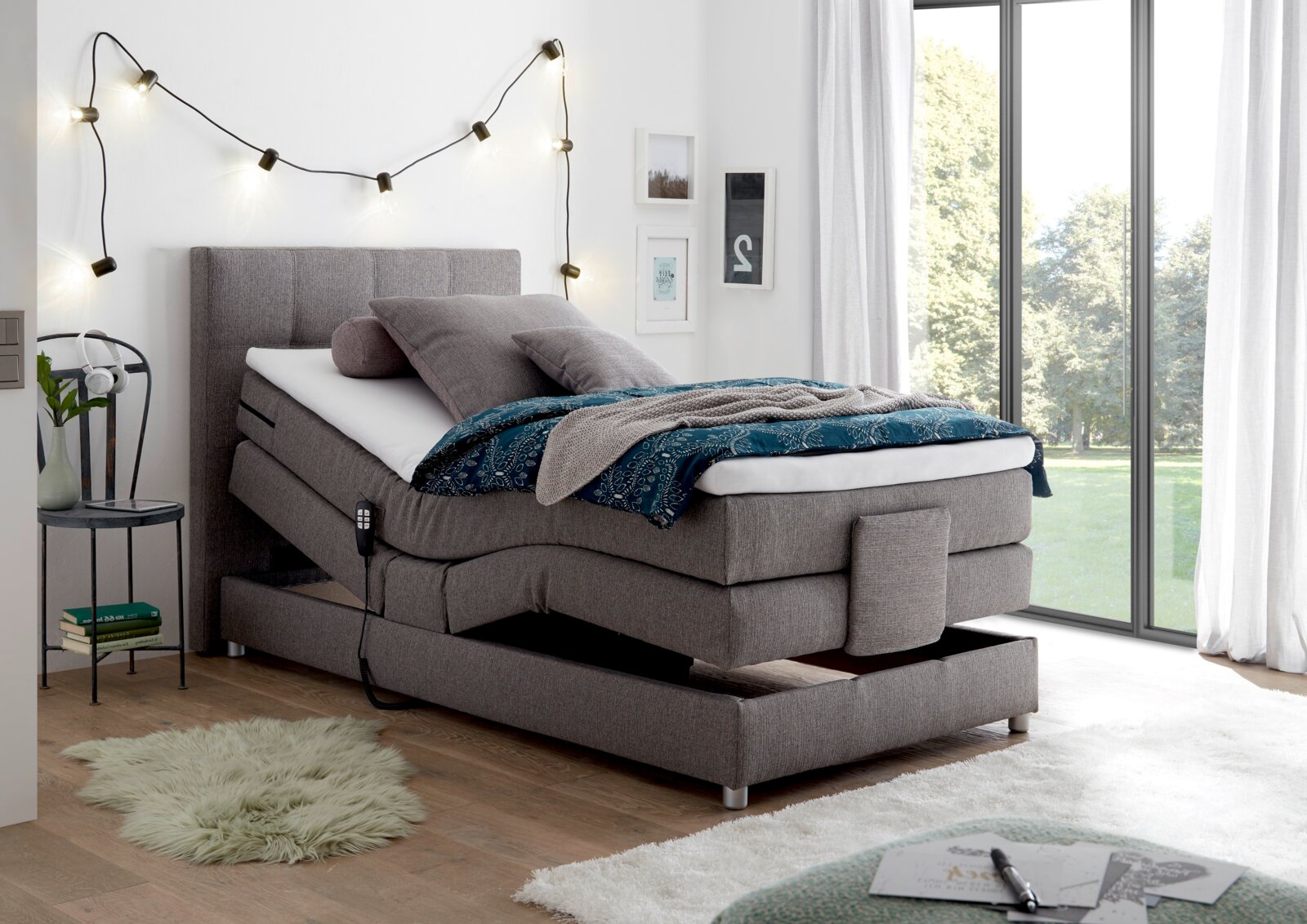 Graues Boxspringbett mit verstellbarem Kopf- und Fußteil, dekoriert mit Kissen und Decken, in einem hellen Raum mit großen Fenstern, seitliche Perspektive.