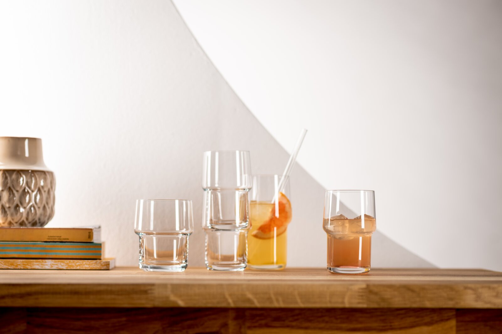 Frontaal aanzicht op een houten plank met meerdere stapelbare transparante glazen; één glas met ijs en amberkleurige drank en een hoog glas met citrusdrank en rietje tegen een lichte muur.