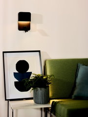 Moderne schwarze Wand- und Bettleuchte BOXER von Lucide, seitliche Perspektive, beleuchtet eine gemütliche Ecke mit grünem Sofa und Kunstwerk.