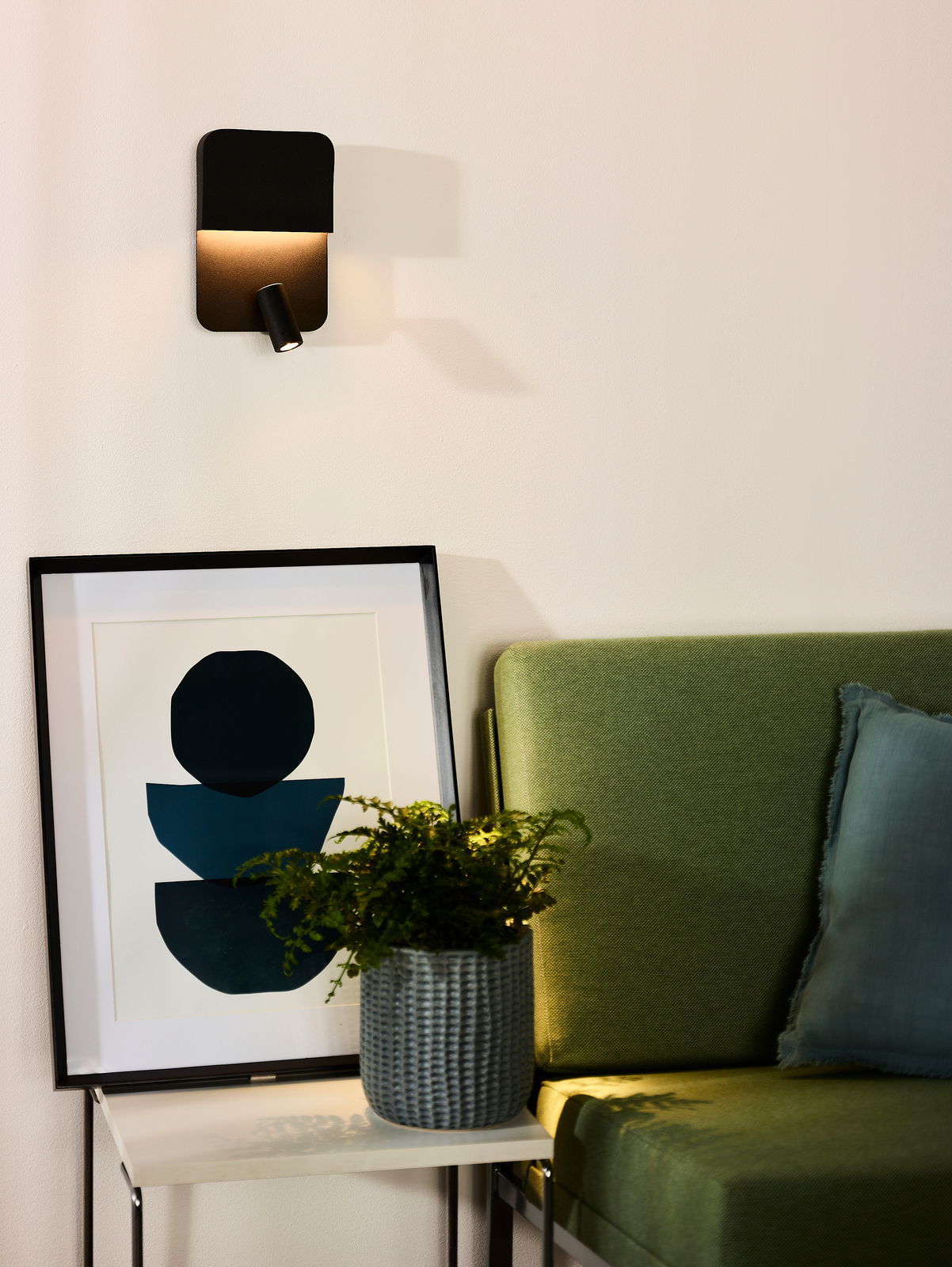 Moderne schwarze Wand- und Bettleuchte BOXER von Lucide, seitliche Perspektive, beleuchtet eine gemütliche Ecke mit grünem Sofa und Kunstwerk.