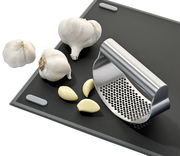 Knoblauchpresse SWING aus Edelstahl auf einem Schneidebrett, umgeben von Knoblauchzehen und -knollen, seitliche Perspektive.