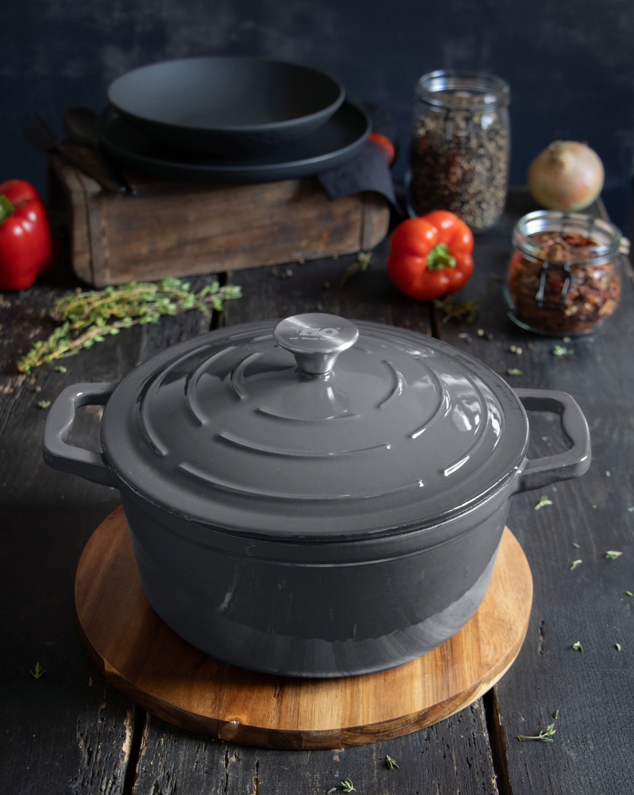 Grauer runder Bräter aus Gusseisen, 26 cm, mit Deckel, auf einem Holzbrett präsentiert, umgeben von Gewürzen und Gemüse, aus einer leicht erhöhten Perspektive fotografiert.