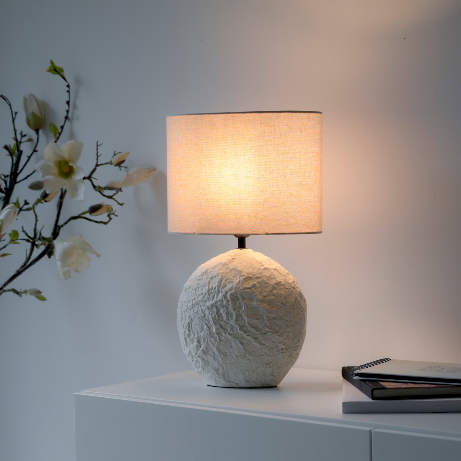 Leuchtende Tischlampe mit weißem, organisch strukturiertem Keramikfuß und zylindrischem Stoffschirm, frontal in einer stilvollen Wohnszene dargestellt