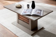 Moderner Couchtisch in Taupe mit Holz-Akzent und breitem Sockel, dekoriert mit offenem Magazin und Keramikvase, fotografiert aus schräger Perspektive von oben (3/4-Ansicht) im Wohnzimmer auf hellem Teppich.
