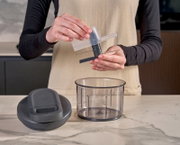 Vooraanzicht op een keukenhakmolen/foodprocessor-accessoire: handen houden het messenblok met transparante spatkap boven een doorzichtige mengkom, met het deksel op het aanrecht.